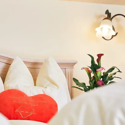 Cozy hotel bed with heart-shaped red pillow and fresh calla lilies beside wall lamp